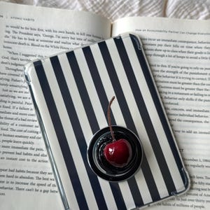 May include: A rectangular phone case featuring a vertical black and white striped pattern. A circular black phone grip is attached to the center, topped with a glossy, realistic red cherry with a brown stem. The case rests on an open book.