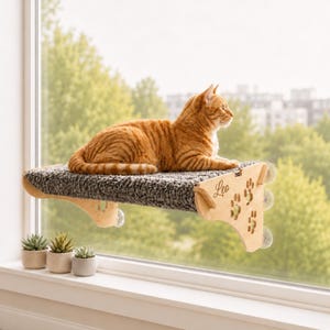 Pode incluir: Um poleiro de janela para gatos em madeira clara com uma superfície de tecido texturizado cinzento-escuro, fixado a uma janela com ventosas. O painel lateral apresenta o nome Leo gravado em cursivo e várias pegadas recortadas.
