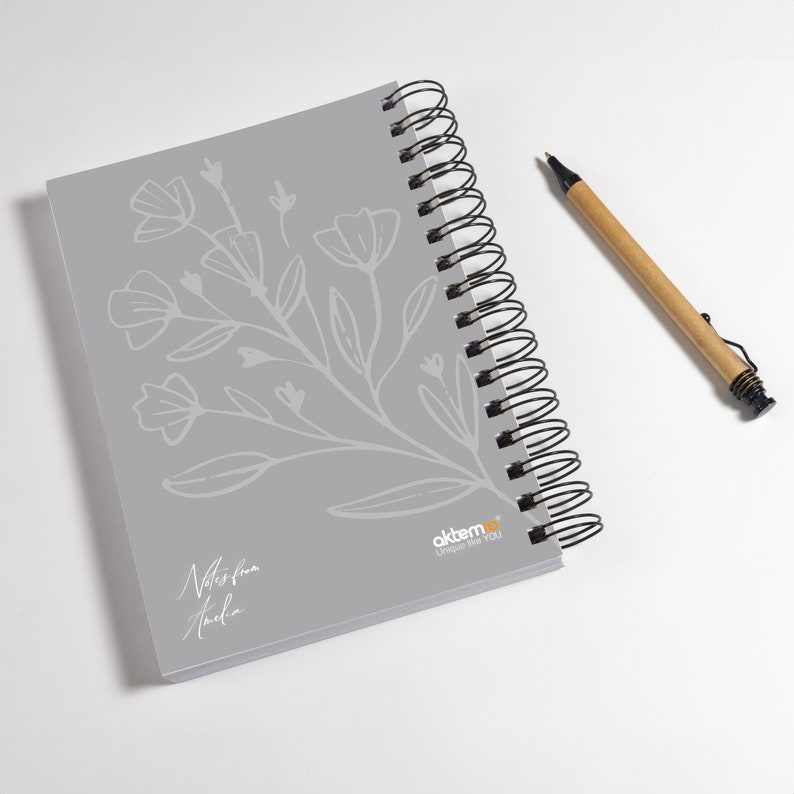 Op de afbeelding: Een grijs spiraalnotitieblok met een bloemenontwerp en de tekst "Notes from Amelia" op de voorzijde. Het notitieblok heeft een zwarte spiraalbinding en de tekst "aktemo&reg; Designed for you" in de rechterbenedenhoek.