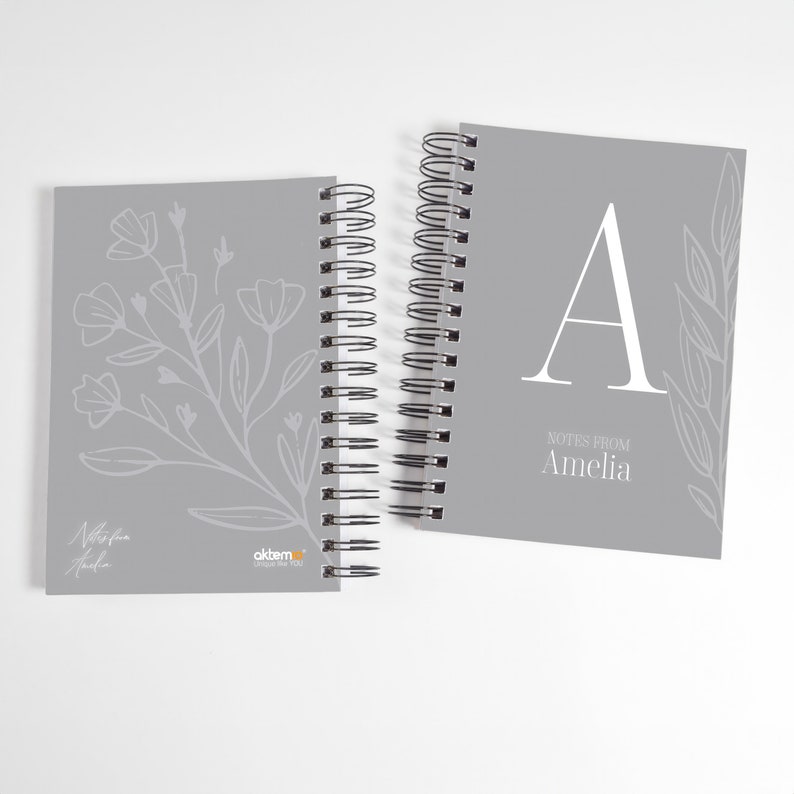 Op de afbeelding: Twee grijze spiraalnotitieblokken met bloemmotieven. Het notitieblok links heeft een bloemmotief en de tekst "Notes from Amelia". Het notitieblok rechts heeft een grote letter "A" en de tekst "Notes from Amelia".