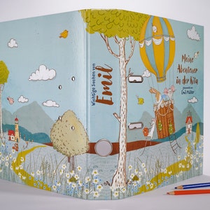 Könnte beinhalten: Ein blauer Ordner mit einer bunten Illustration eines Heißluftballons, eines Kaninchens und einer Maus. Der Ordner hat den Text "Wichtige Sachen von Emil" und "Meine Abenteuer in der Kita" auf dem Cover.