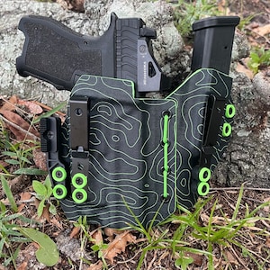 May include: A black and green patterned holster with a black handgun and magazine inside. The holster has a black clip and green accents.