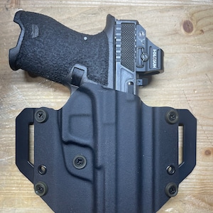 May include: A black handgun with a red dot sight attached to the top. The gun is in a black holster with a belt loop.