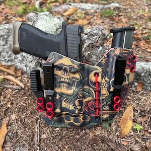 May include: A black and tan leather holster with a skull and floral print design. The holster has red stitching and red snaps. A black handgun is in the holster.