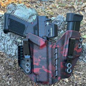 May include: A black handgun with a red dot sight and a black magazine is placed in a red and black camouflage holster. The holster has two black clips and gray lacing. The gun is resting on a rock.