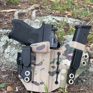 May include: A black and tan camouflage holster with a black gun and magazine inside. The holster has a black clip and a black strap with the word "MULTICAM" printed on it. The holster is sitting on a bed of brown leaves and green grass.