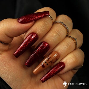 Pode incluir: Unhas compridas de cor vermelha com glitter, com uma &uacute;nica unha de destaque com um design de rena marrom.