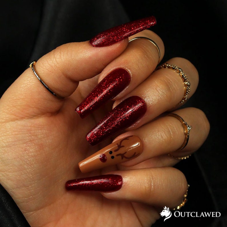 Pode incluir: Unhas compridas de glitter vermelho com uma &uacute;nica unha de destaque com um design de rena marrom. A rena tem um nariz vermelho e chifres.