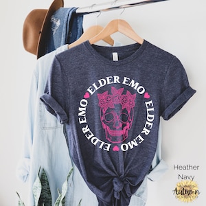 May include: A heather navy blue t-shirt with a white circle graphic featuring a skull with a floral crown and the text "Elder Emo" repeated around the skull. The text is in a pink font.