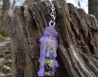 May include: A small glass bottle filled with dried herbs and flowers, encased in a purple wax coating. The bottle is attached to a silver chain, suggesting it is a necklace or charm.