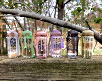 May include: Six small glass jars with cork tops filled with various herbs and spices. The jars are sealed with wax and have different colored wax drippings. The jars are arranged in a row on a wooden surface.