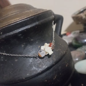 May include: A silver chain necklace with three beads. The beads are a clear quartz crystal, a white bone bead, and a red and orange agate bead.
