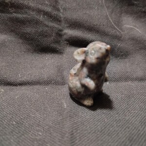May include: A small, stone-carved figurine of a mouse. The mouse is gray and brown with visible details, including ears, a nose, and small feet. The figurine is set against a dark, textured background.