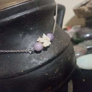 May include: A silver chain necklace with three beads. The beads are purple and white. The necklace is on a dark brown surface.
