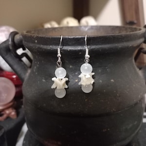 May include: A pair of silver earrings with white beads and bone-shaped charms. The earrings are hanging from a dark background.