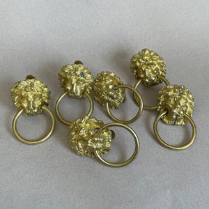 May include: Six gold lion head drawer pulls with ring handles. The lion heads are detailed and have a textured finish.