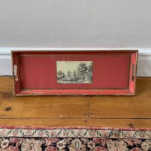 May include: A red wooden tray with a gold trim and a black and white print of people on horseback. The tray is on a wooden floor with a patterned rug in the foreground.