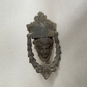May include: A vintage cast iron door knocker featuring a detailed design of a face with a crown. The knocker is dark brown in colour and has a worn, aged appearance.
