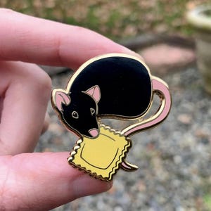 May include: A black enamel pin of a rat holding a yellow ravioli. The rat has pink ears and a pink tail.
