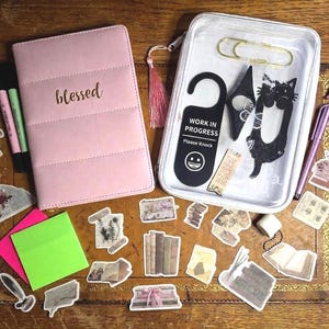 May include: A collection of stationery items on a wooden surface. Includes a pink notebook with "blessed" in gold lettering, markers, stickers, a clear tray with a paperclip and cat-shaped cutout, and a "Work in Progress" door hanger.