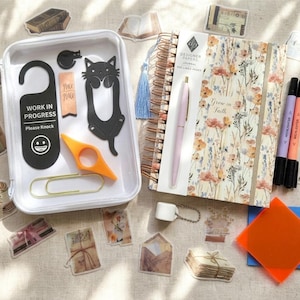 May include: A collection of stationery items including a floral journal, pen, highlighters, stickers, and a clear case with a cat-shaped bookmark, door hanger with "Work in Progress" text, and an orange page holder.
