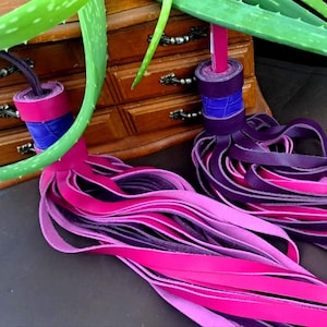 May include: Two decorative tassels with layered leather strips in shades of pink and purple. The tassels have rolled tops with blue and purple accents. A wooden chest of drawers and aloe vera plant are in the background.