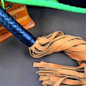 May include: A black leather whip with a braided handle and tan leather fringe. The handle is a dark blue color. The whip is lying on a dark surface.