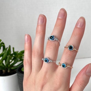 Pode incluir: Tr&ecirc;s an&eacute;is de prata e ouro com pingentes de olho azul mau. Os an&eacute;is s&atilde;o usados nos dedos de uma pessoa.