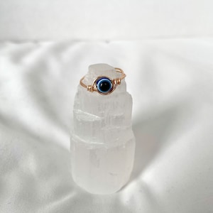 Pode incluir: Anel de fio de ouro com um amuleto de olho azul. O anel &eacute; exibido em um cristal de selenita branco.