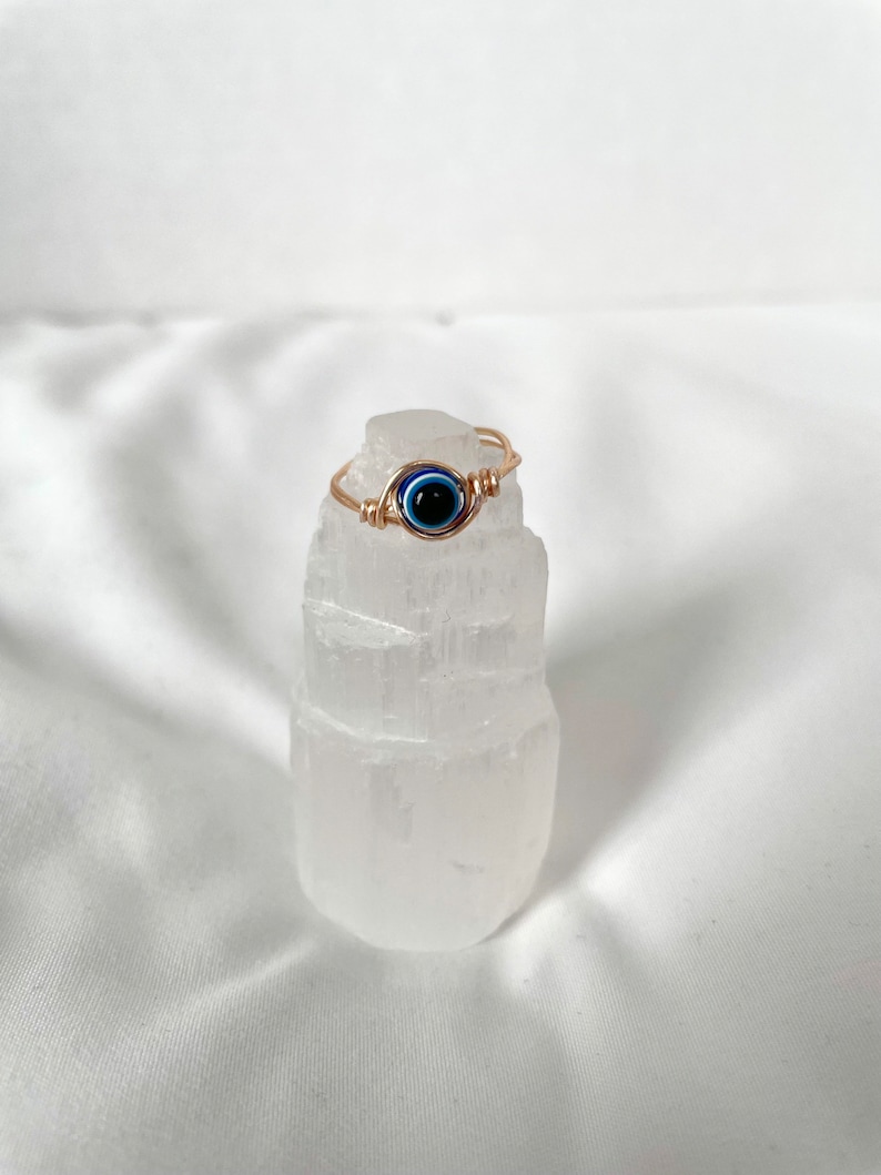 Pode incluir: Anel de fio de ouro com um amuleto de olho azul. O anel &eacute; exibido em um cristal de selenita branco.