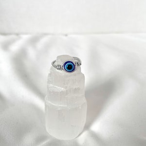 Pode incluir: Um anel de prata com um amuleto de olho turco azul. O anel &eacute; apresentado em um cristal de selenita branco.