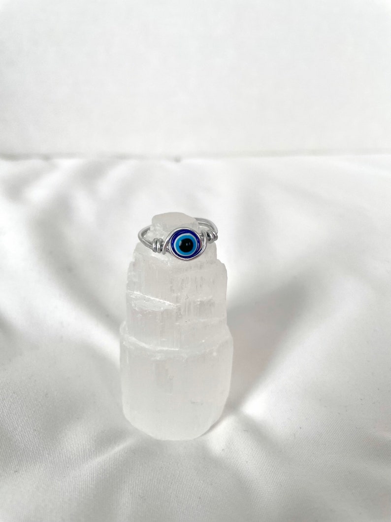 Pode incluir: Um anel de prata com um amuleto de olho turco azul. O anel &eacute; apresentado em um cristal de selenita branco.