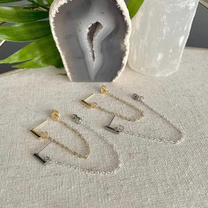 Peut inclure: Trois paires de boucles d'oreilles à chaîne dorées, argentées et noires. Chaque boucle d'oreille a une petite barre et une chaîne qui pend du lobe de l'oreille.