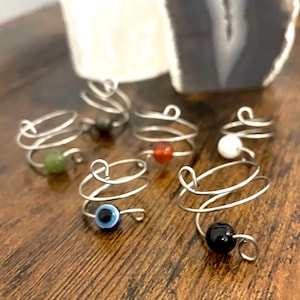 May include: A collection of six silver-toned wire rings, each featuring a single bead. The beads include a green stone, a dark grey stone, a red stone, a white stone, a blue and black evil eye charm, and a solid black stone.