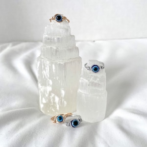 Pode incluir: Tr&ecirc;s an&eacute;is envoltos em fio de prata e ouro com pingentes de olho azul mau. Os an&eacute;is s&atilde;o exibidos em torres de cristal de selenite branca.