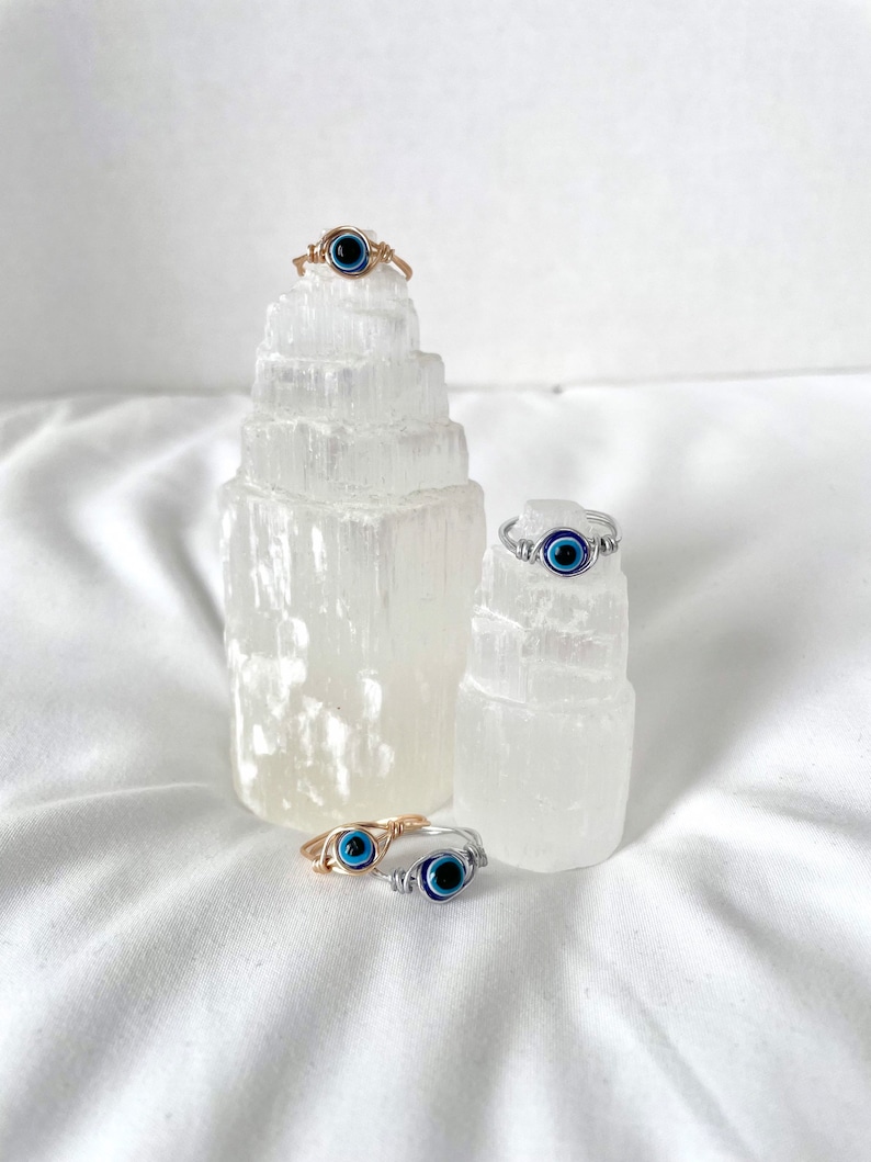 Pode incluir: Tr&ecirc;s an&eacute;is envoltos em fio de prata e ouro com pingentes de olho azul mau. Os an&eacute;is s&atilde;o exibidos em torres de cristal de selenite branca.