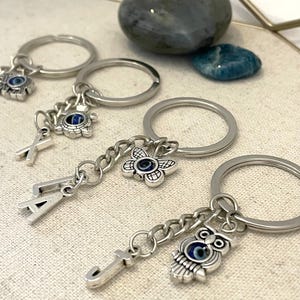 May include: Silver keyrings with charms, including an owl, a flower, and letters. The charms feature blue and white evil eye designs. The keyrings are displayed on a textured, off-white surface, with two stones in the background.
