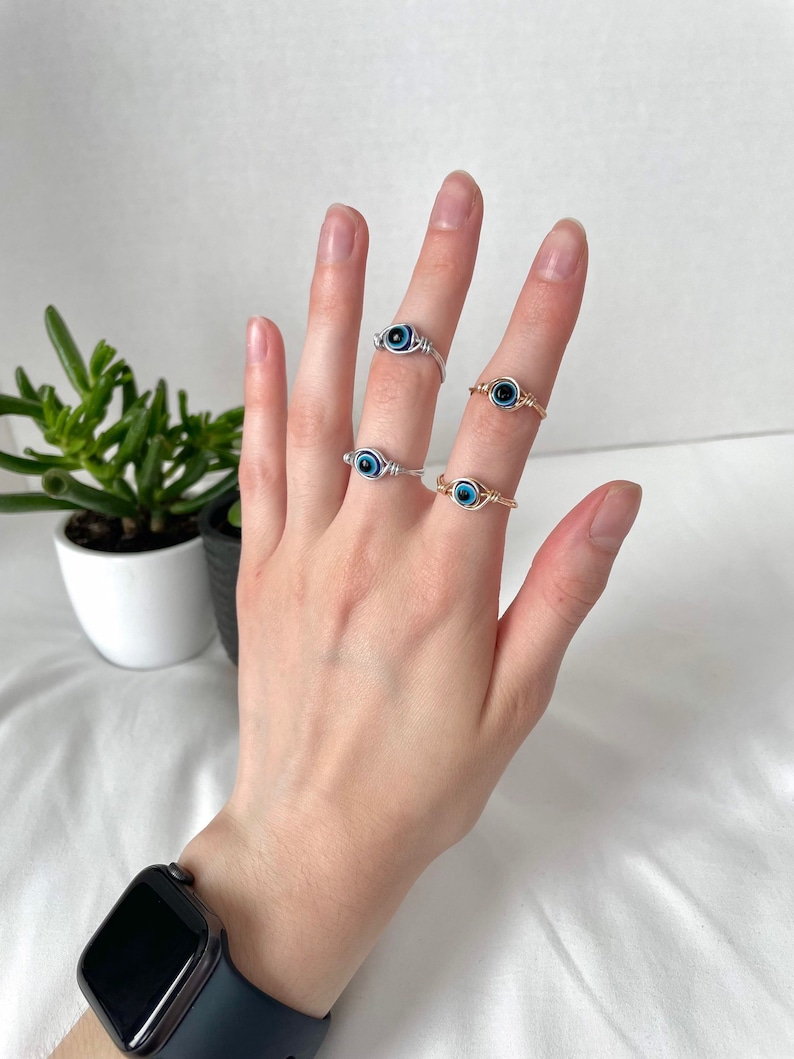 Pode incluir: Tr&ecirc;s an&eacute;is de prata e ouro com pingentes de olho azul mau. Os an&eacute;is s&atilde;o usados nos dedos de uma pessoa.