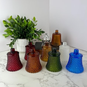 Puede incluir: Una colección de coloridos candelabros de vidrio en varios tonos, incluyendo rojo, ámbar, verde, blanco y azul. Cada portavelas tiene un patrón texturizado, similar a un clavo, y una tapa a juego. Una planta decorativa en una maceta blanca está en el fondo.