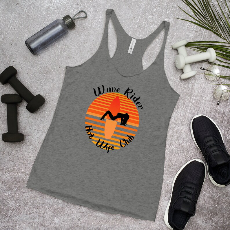 May include: Gray racerback tank top with a graphic featuring a surfer on a surfboard against an orange sunset. The text reads "Wave Rider Hot Wife Club". The tank top is surrounded by workout equipment and athletic shoes.