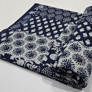 Puede incluir: Un textil plegado con fondo azul marino y estampado floral blanco. El patrón incluye diversas formas, como puntos, diseños florales y otras formas orgánicas. La tela está parcialmente enrollada, revelando el patrón.