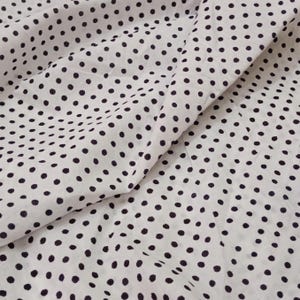 May include: White fabric with a repeating pattern of dark navy blue polka dots. The fabric is slightly wrinkled, creating shadows and depth. The polka dots are evenly spaced across the material.