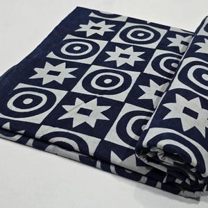 Puede incluir: Textil plegado y enrollado con un patrón geométrico repetitivo. El diseño presenta formas blancas, incluyendo estrellas, círculos y cuadrados, sobre un fondo azul marino. La tela está doblada y enrollada.