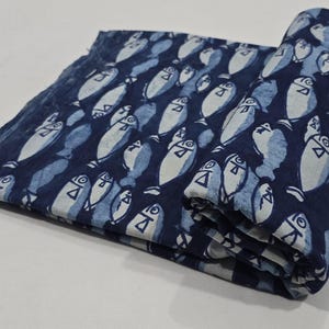Puede incluir: Tela enrollada con un patrón repetitivo de peces blancos y azul claro sobre un fondo azul oscuro. La tela está doblada, revelando el patrón y la textura. Ideal para manualidades o decoración del hogar.