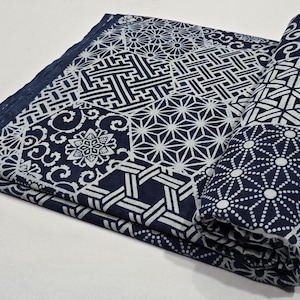 Puede incluir: Una tela plegada y enrollada de color azul marino y blanco. La tela presenta diseños geométricos y florales en blanco sobre un fondo azul oscuro. La parte enrollada muestra un patrón de puntos repetidos.