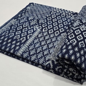 May include: Folded indigo blue fabric with white geometric patterns. The fabric features a repeating diamond and floral design, with a rolled section on the right. The fabric is likely used for sewing or crafting.