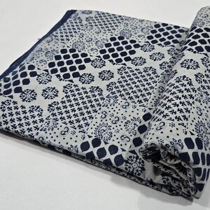 May include: A folded textile with a patchwork design in white and navy blue. The fabric displays various geometric and floral patterns. One side is rolled up, showing the pattern and texture. The textile is likely a blanket or throw.