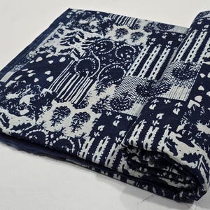 May include: A folded indigo and white patterned textile. The fabric features a repeating design of geometric shapes, trees, and abstract patterns. The textile is partially rolled, revealing the intricate design. The color palette is a deep indigo blue and white.