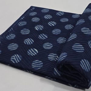 Op de afbeelding: Gerolde marineblauwe stof met een herhalend patroon van witte gestreepte cirkels. De stof is gevouwen en gerold, waardoor het ontwerp wordt getoond. De achtergrond is een effen wit oppervlak.
