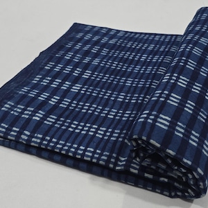 May include: A folded textile with a blue and white plaid pattern. The fabric is a deep indigo blue with a grid of white lines creating the plaid design. The textile is partially rolled, showing the pattern.